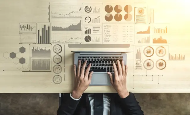 Person in business attire typing on a laptop at a desk, with digital charts, graphs, and data visualizations projected on the tabletop, symbolizing business analytics and big data technology