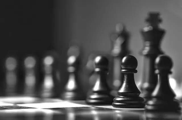 Close-up black and white photo of black chess pieces on a chessboard, with pawns in focus and the king blurred in the background.